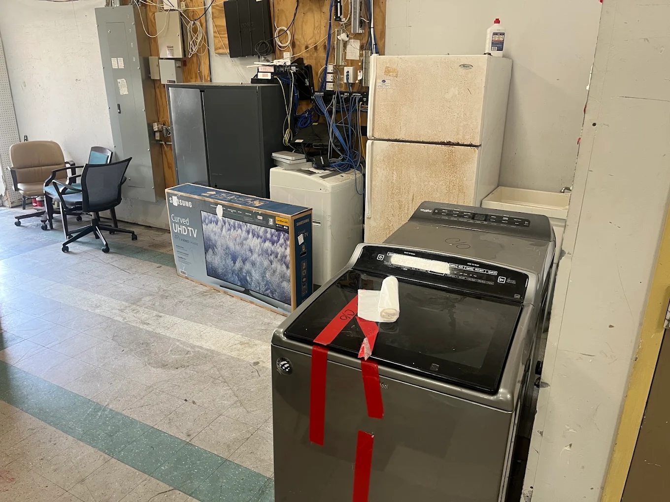 Washers, a dryer, a refrigerator, office chairs and a boxed TV staged inside a warehouse for appliance removal