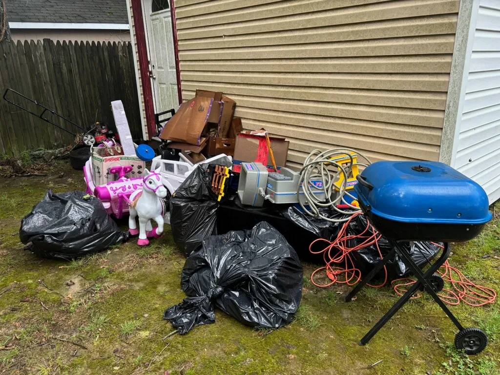 Backyard pile of kids' toys, a grill, hoses and bagged junk staged beside a shed for removal