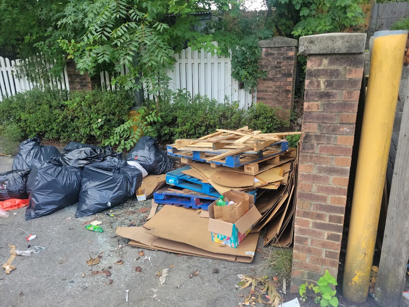 Alley pile of pallets, flattened cardboard and bagged trash behind a rowhouse fence
